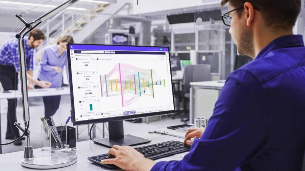 A person in a blue shirt is working on a computer displaying a data analysis dashboard, with two colleagues discussing in the background.
