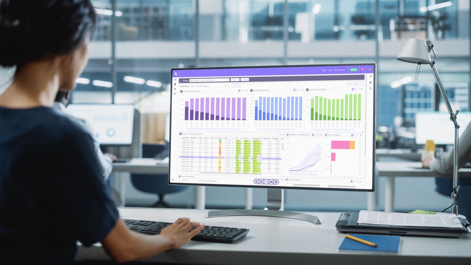 A person in a blue shirt is analyzing forecasting data on a computer screen, with two colleagues collaborating in the background.