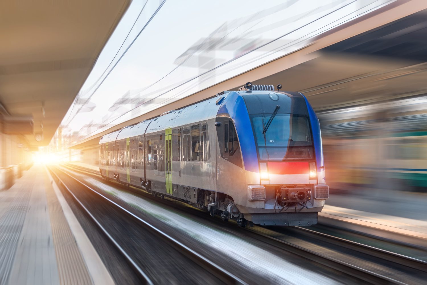 A sleek blue and silver passenger train speeds through a modern train station, with a blurred background indicating motion.